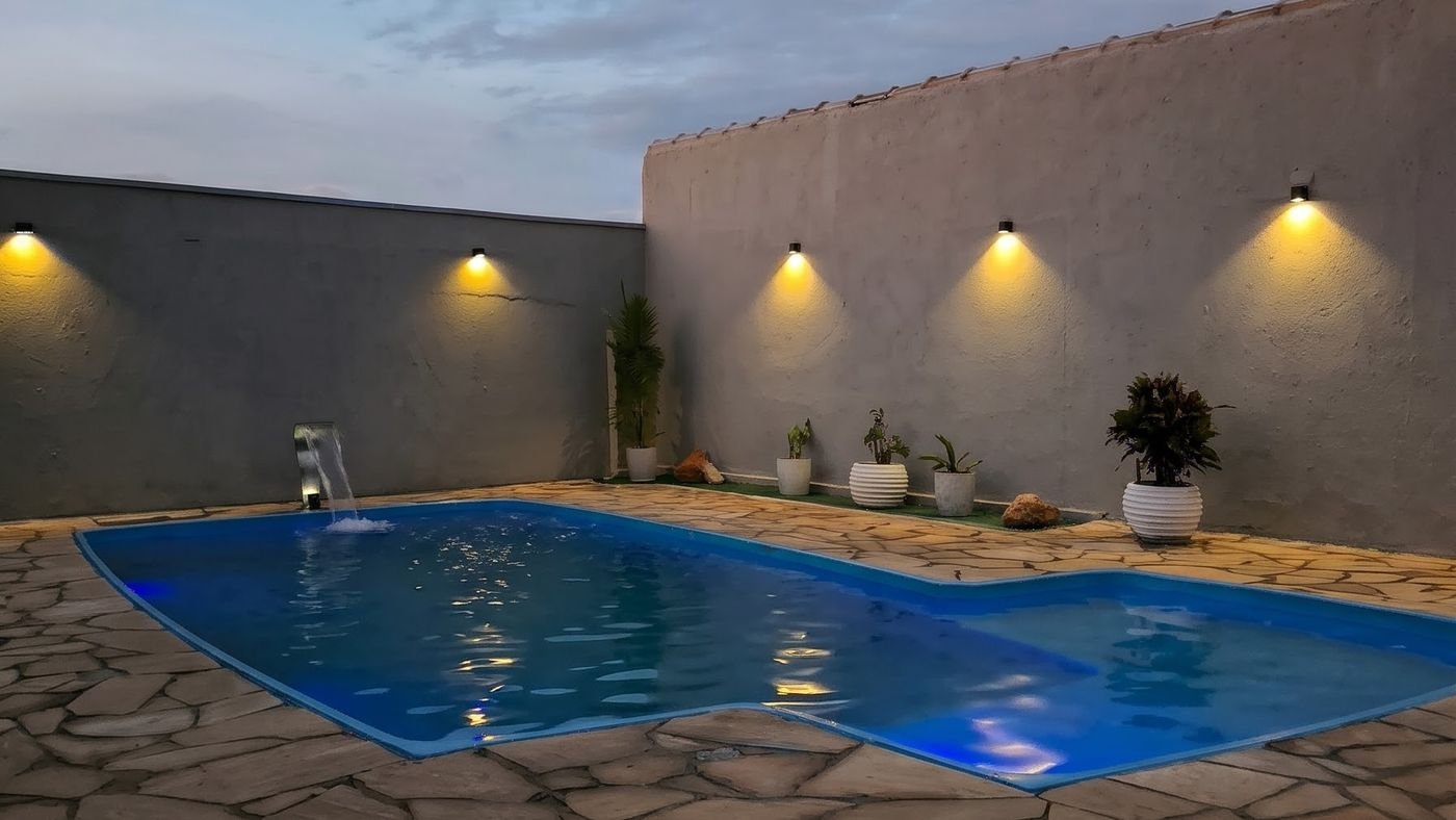Piscina iluminada ao entardecer com arandelas de luz quente acesas no muro, projeto Domus em Sete Lagoas